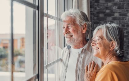 Happy bonding loving middle aged senior retired couple standing near window, looking in distance, recollecting good memories or planning common future, enjoying peaceful moment together at home.